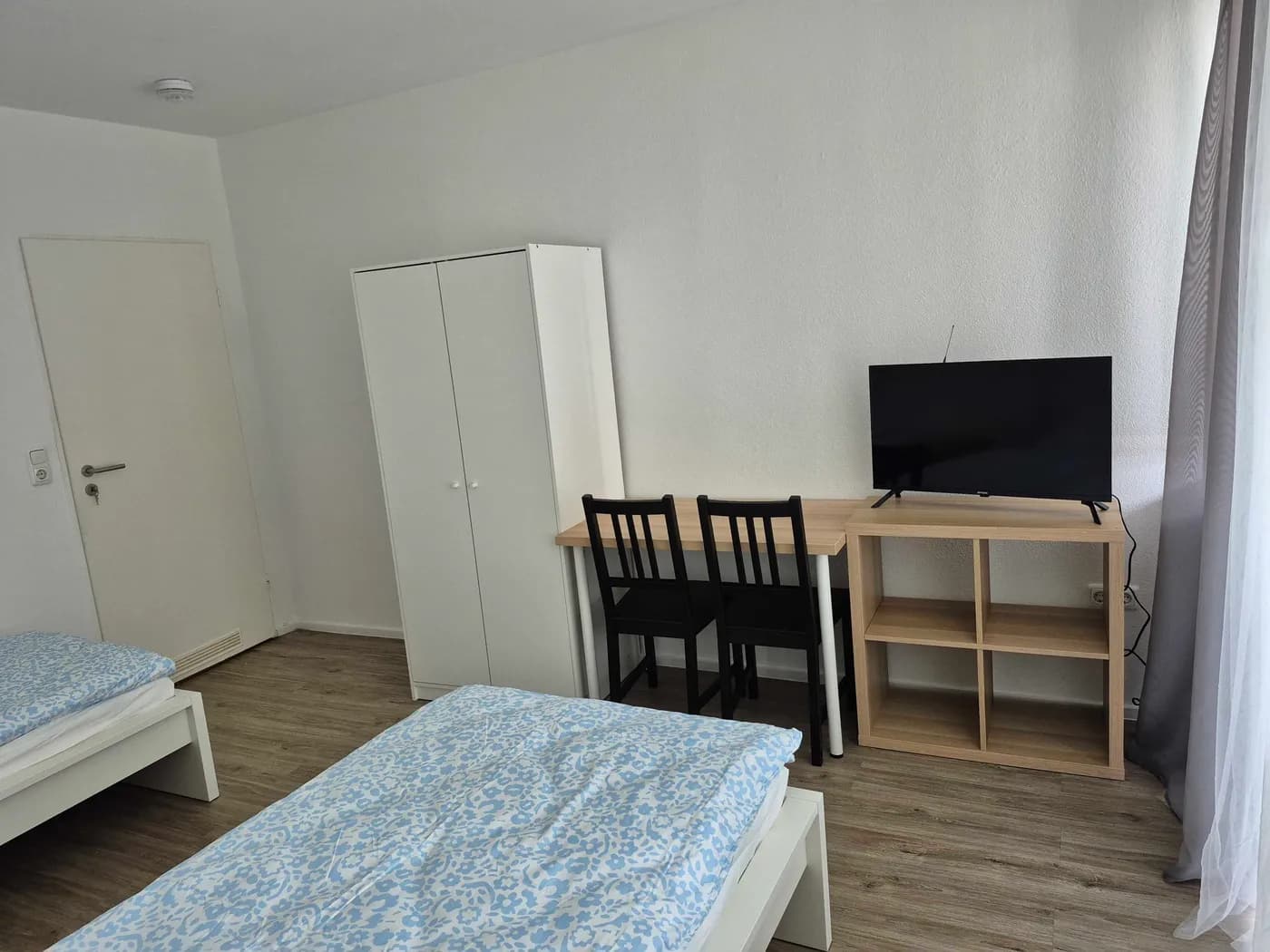 Helles Schlafzimmer einer Monteurwohnung in Offenbach mit zwei getrennten Einzelbetten, Kleiderschrank, Schreibtisch mit Stühlen und Fernseher