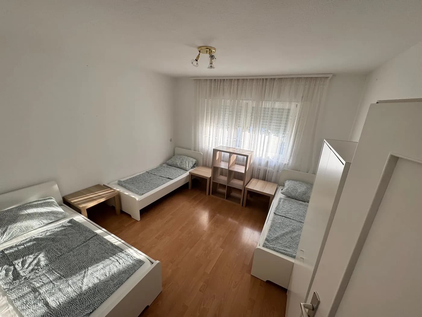 Helles Schlafzimmer einer Monteurwohnung in Dreieich mit drei getrennten Einzelbetten, Regal und Fenster mit Gardine auf Laminatboden