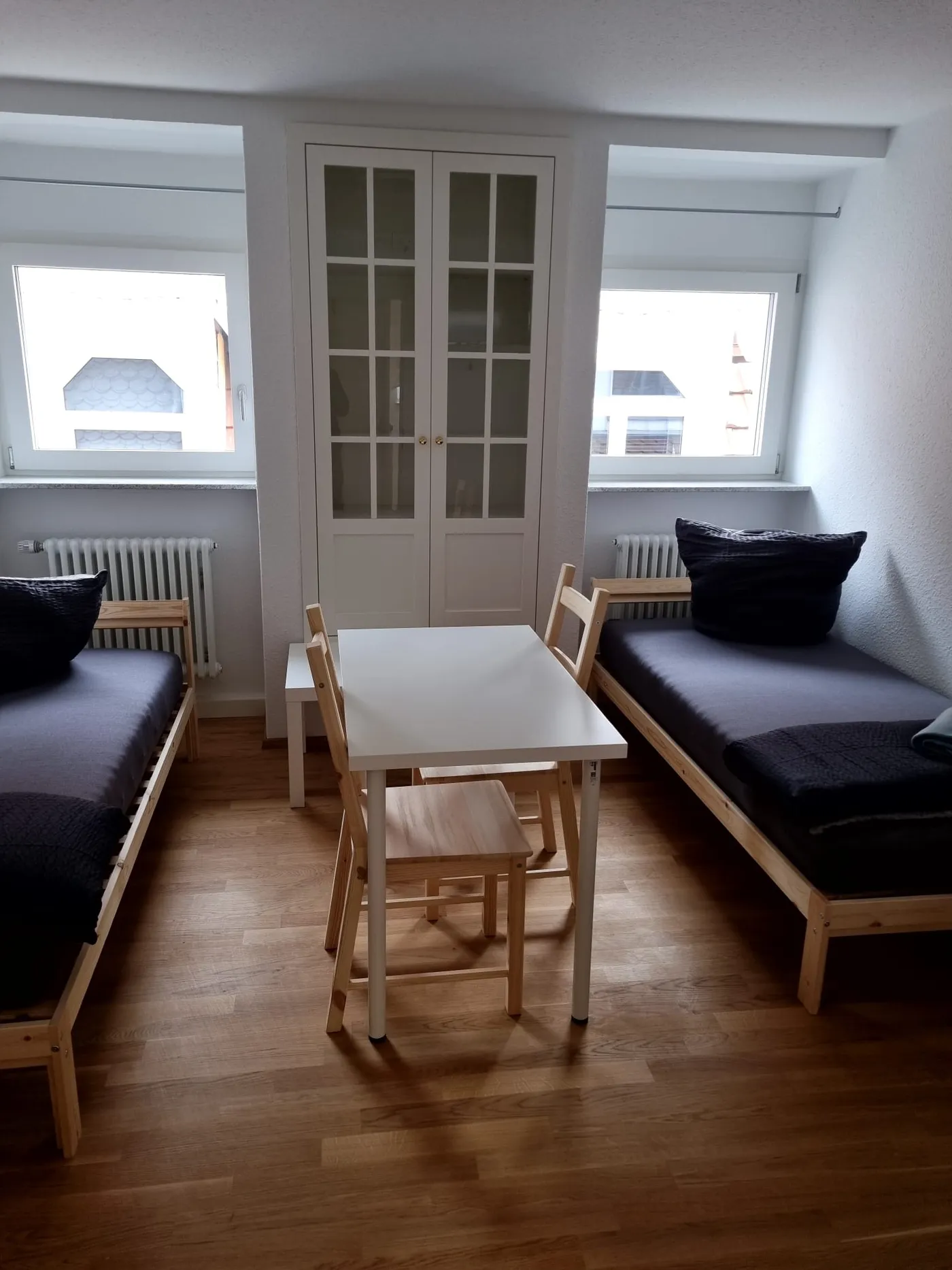 Helles Schlafzimmer einer Monteurwohnung in Friedrichshafen mit zwei getrennten Einzelbetten, Esstisch und Vitrinenschrank auf Eichenparkett